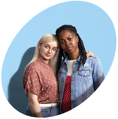 A young White woman, wearing a pink blouse and red glasses, puts her arm around a young Black woman, wearing a denim jacket. They are both standing proud.